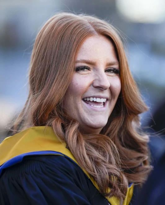 Close-up of a smiling graduate with red hair wearing a black gown and gold sash. Close-up of a smiling graduate with red hair wearing a black gown and gold sash.